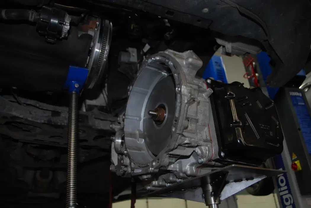 An automatic gearbox being carefully lowered from a vehicle using a hydraulic transmission jack in a workshop, showing the exposed torque converter housing and transmission assembly during removal.
