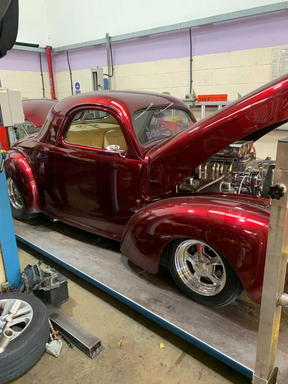 Custom-built burgundy Willys Coupe hot rod with polished chrome wheels on a workshop ramp
